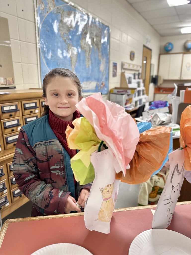 Alt text: A smiling student stands in a classroom beside colorful tissue-paper flower creations displayed on a table, with a world map visible in the background.