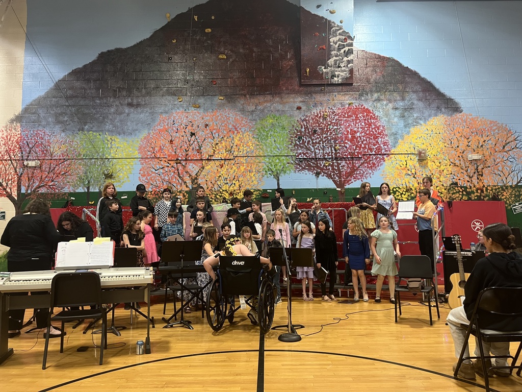 Alt text: Students stand on risers in a gymnasium preparing for a performance, with musical instruments and a large mural of trees and mountains in the background.