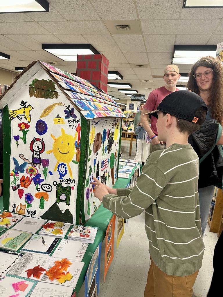 Alt text: A student explores a large decorated cardboard house covered in colorful drawings and artwork while families browse displays in the background.