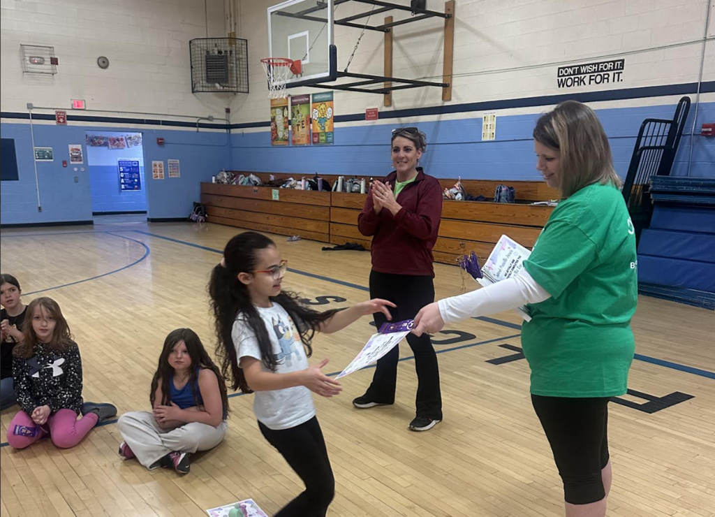 A student receives a certificate and ribbon from a staff member during a school recognition event in the gym while classmates sit and watch nearby.