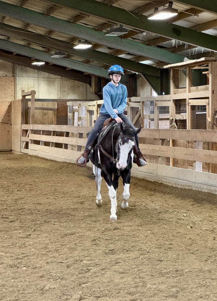 A student rides a horse in an indoor riding arena while wearing a helmet and holding the reins.