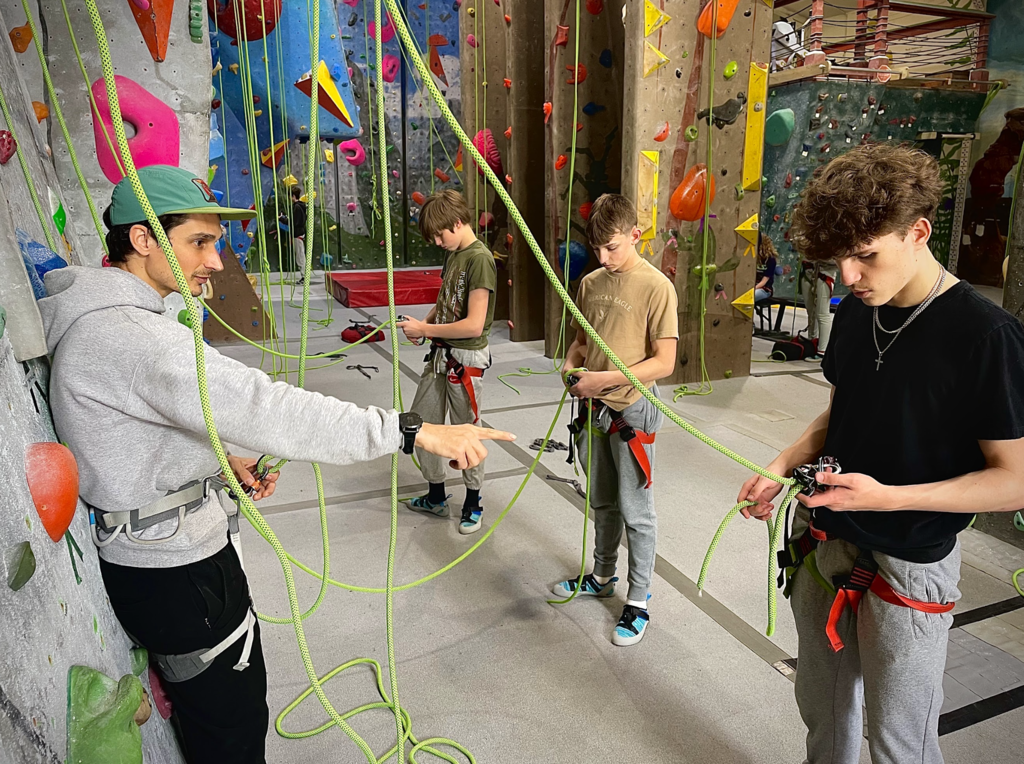 Students at an indoor climbing gym learn rope and harness techniques while an instructor points and provides guidance.