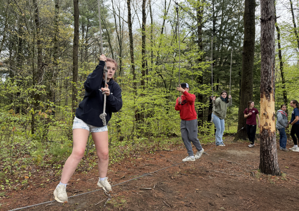Students participate in an outdoor ropes challenge in a wooded area, balancing and holding ropes while navigating the activity course.