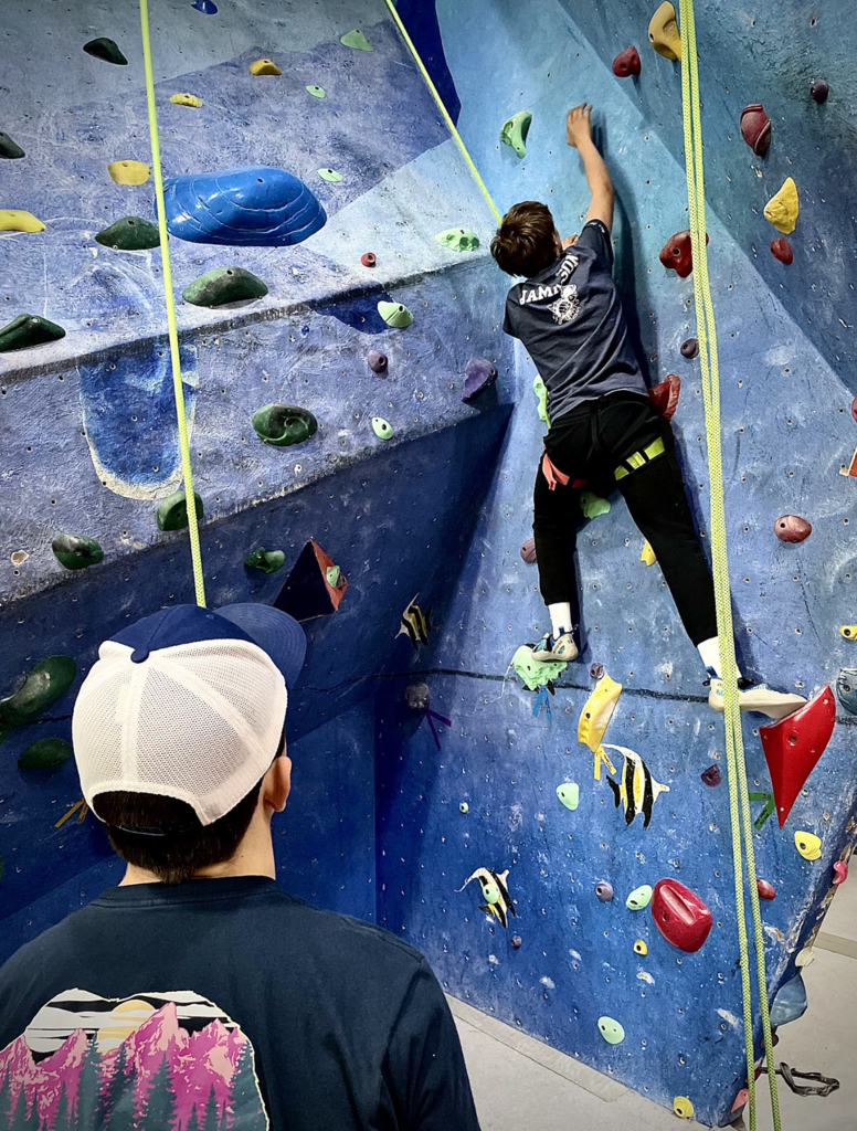 A student climbs a blue indoor rock wall while another student watches from below.