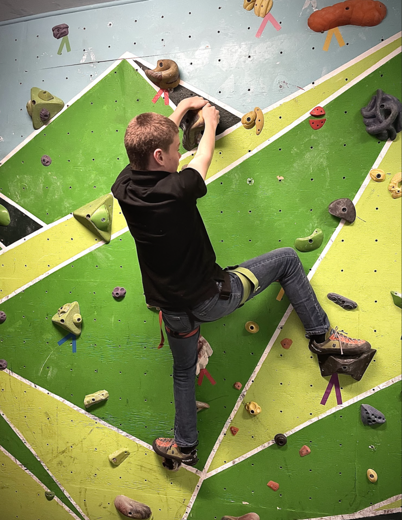 A student climbs a bright green indoor rock wall while wearing a harness and climbing shoes.