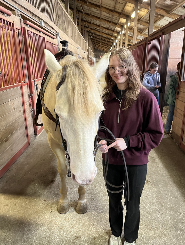 A student stands beside a light-colored horse in a stable aisle holding the reins and smiling for a photo.