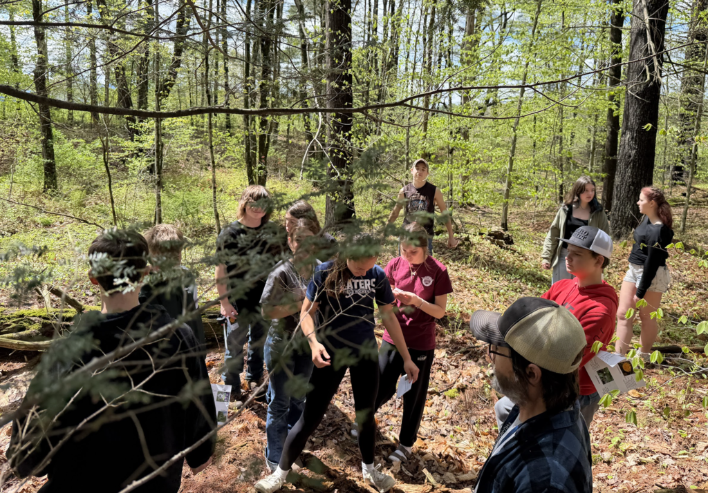 group of students stands together in a wooded area during an outdoor learning activity, listening and exploring the surroundings.