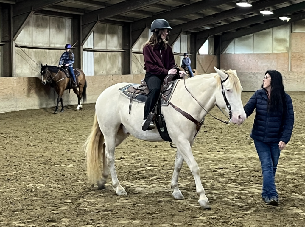 A student rides a white horse in an indoor arena while an instructor walks beside them and other riders practice in the background.