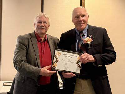 Two men stand side by side indoors holding a framed certificate and smiling for a photo during an awards presentation. One man is presenting the certificate while the recipient wears a boutonniere.