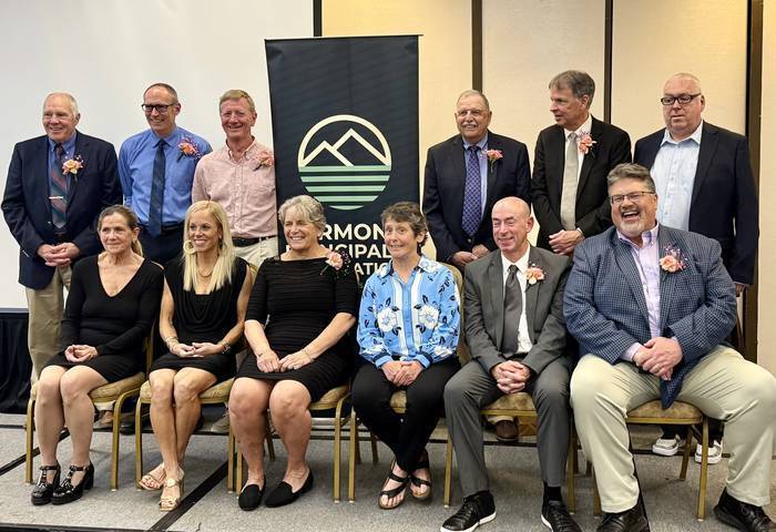 A group of award recipients and honorees pose for a group photo in front of a banner at an indoor recognition event. Several individuals are seated in the front row while others stand behind them, many wearing boutonnieres or corsages.