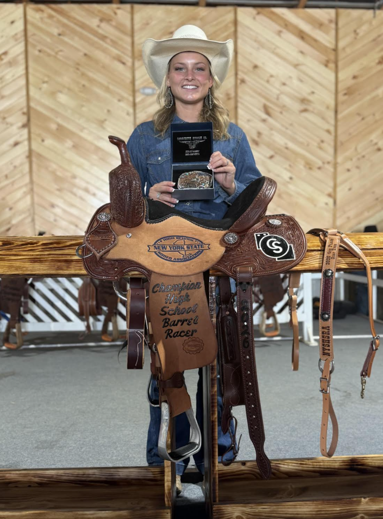 A smiling student wearing a white cowboy hat and denim shirt stands behind a decorated championship saddle while holding an award plaque and belt buckle display. The saddle is engraved with “Champion High School Barrel Racer,” recognizing a rodeo accomplishment. The photo was taken indoors against a wooden wall backdrop.