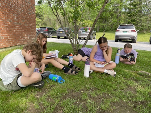 Five students sit and lie on the grass under a tree reading books and writing in notebooks during outdoor learning time.