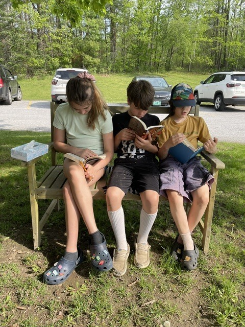 Three students sit together on a wooden bench outside, each focused on reading a book in the shade of a tree.