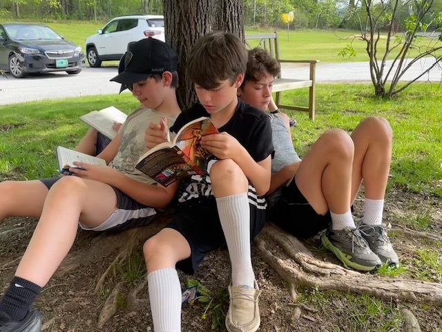 Three students sit under a tree with their backs against the trunk, quietly reading books outdoors near a grassy area and parking lot.