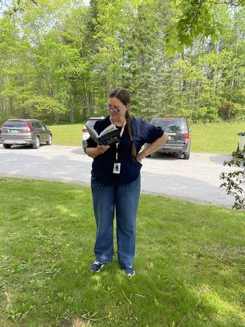 A staff member stands on the grass outdoors reading a book while wearing glasses and a lanyard, with trees and parked cars in the background.