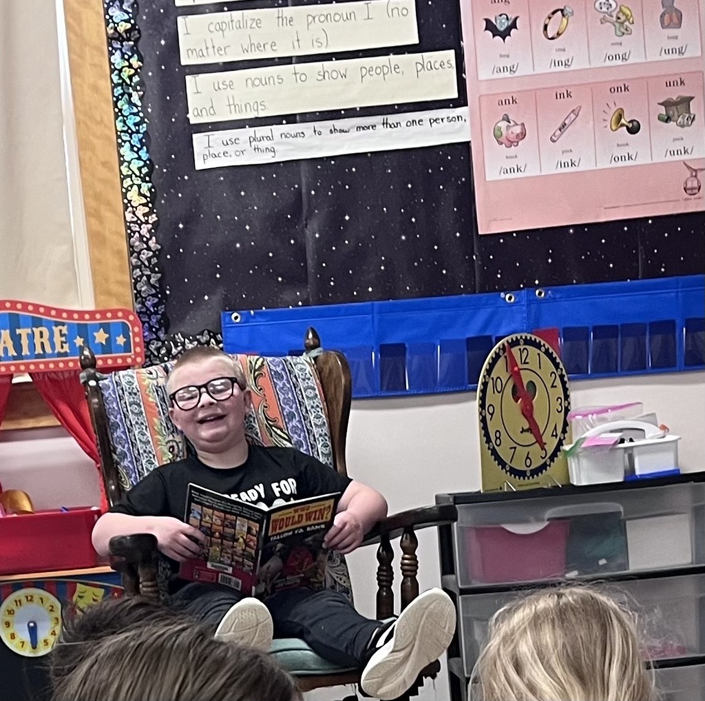 A young boy with glasses sits comfortably in a wooden rocking chair, smiling brightly while holding open a book titled "Who Would Win?". He wears a black T-shirt and has his feet propped up. In the background, classroom decorations are visible, including a poster about grammar rules, a blue pocket chart, and a large toy clock showing the time. The tops of two other children's heads are visible in the foreground.