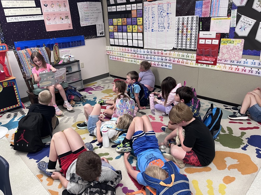 A young girl with long brown hair sits in a wooden rocking chair in a colorful classroom, reading a picture book aloud to a group of children. The children are sitting on a patterned rug decorated with multi-colored handprints, some listening attentively while others lean back or sit with backpacks on. The classroom background features educational posters, including a large calendar, number charts, and grammar rules.