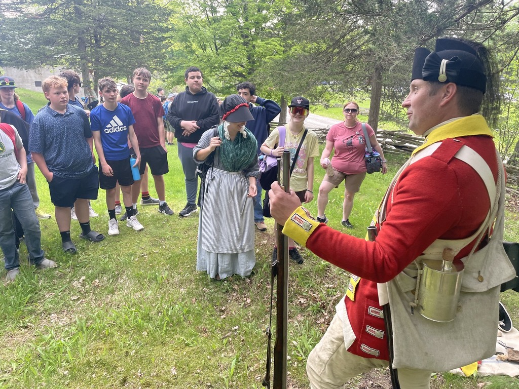An 18th-century reenactor dressed in a bright red military uniform speaks with a group of 8th grade students gathered outdoors on a grassy area surrounded by trees during a visit to Mount Independence.