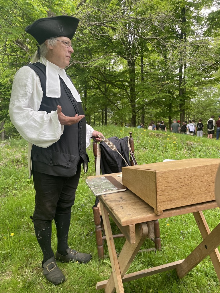 A historical interpreter dressed in black colonial-era clothing stands beside a wooden table and speaks to students in a grassy wooded area during a Mount Independence presentation.