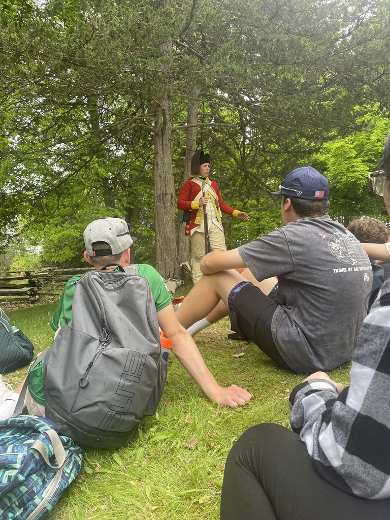 Students sit on the grass listening to a Revolutionary War reenactor dressed in a red military coat speaking beneath tall trees.