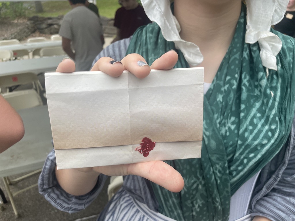 A student dressed in colonial-style clothing holds up a folded paper letter sealed with red wax while standing under a tented area.