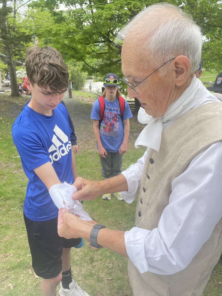 A historical interpreter demonstrates a cloth bandage or fabric activity with a student while other students watch nearby outdoors.