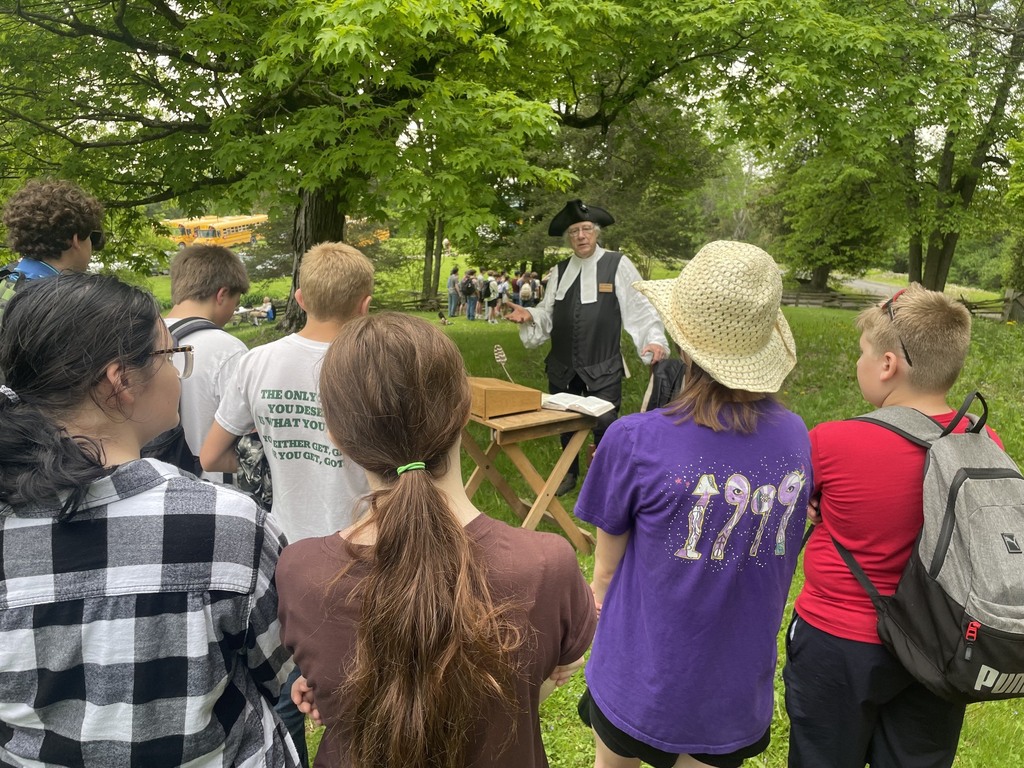 Students stand together outdoors listening to a historical interpreter dressed in colonial clothing speaking beside a wooden table under large green trees at Mount Independence.