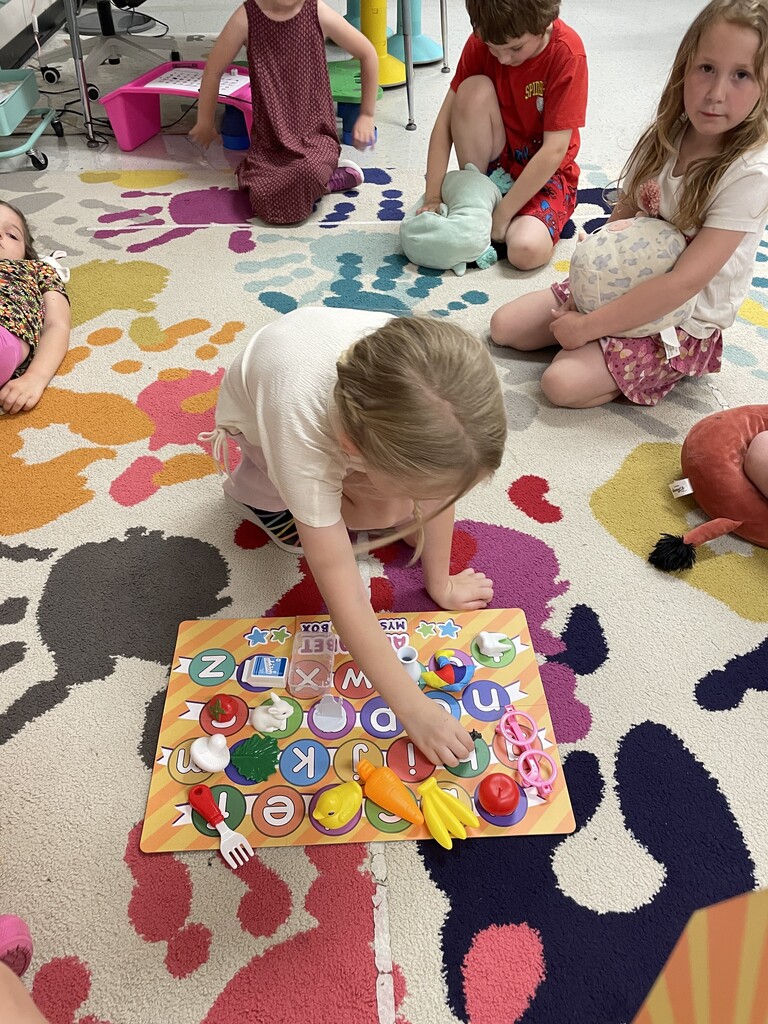 A high-angle shot of a young girl with blonde hair tied in a braid, kneeling on a brightly patterned classroom rug. She is reaching down to place a small toy miniature onto a colorful alphabet matching board spread out on the floor. Several other children are sitting in a circle around her, watching or holding plush toys.