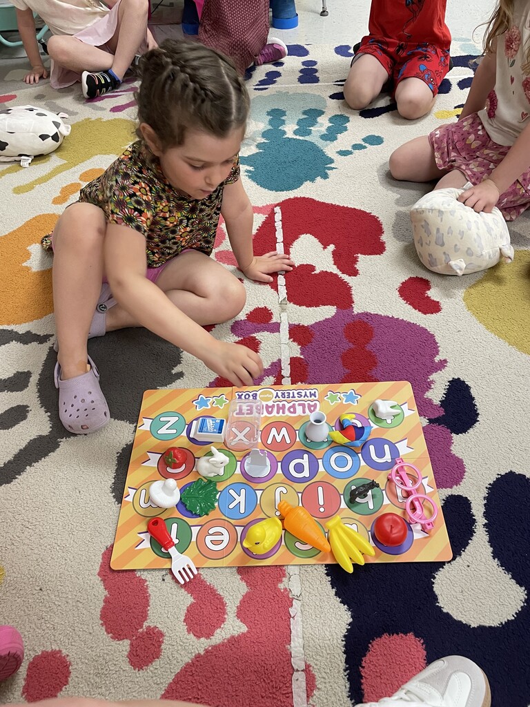 : A young girl with braided hair and a floral top sits on a multi-colored rug, looking down at an alphabet matching game board. She is using her hand to place small plastic objects—including a miniature carrot, banana, duck, and glasses—onto their corresponding letters on the board, while other children watch from the surrounding circle.