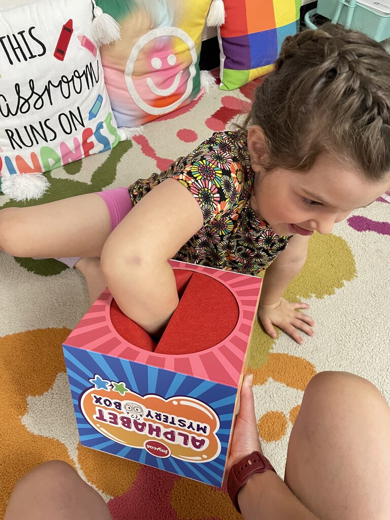 A close-up shot of a young girl with braided hair wearing a floral shirt, sitting on a colorful classroom rug. She is smiling as she reaches her arm deep into the soft, red felt opening of a colorful toy cube labeled "Alphabet Mystery Box." Another person's hands are visible at the bottom, holding the box steady for her.