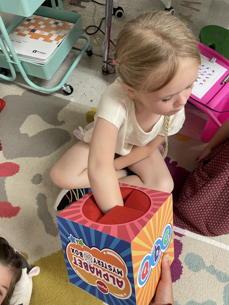 : A young blonde girl with a side braid sits cross-legged on a classroom rug, looking focused as she reaches her arm into the top of a vibrant "Alphabet Mystery Box." The colorful, striped cardboard toy box is being held from below by a teacher or classmate.