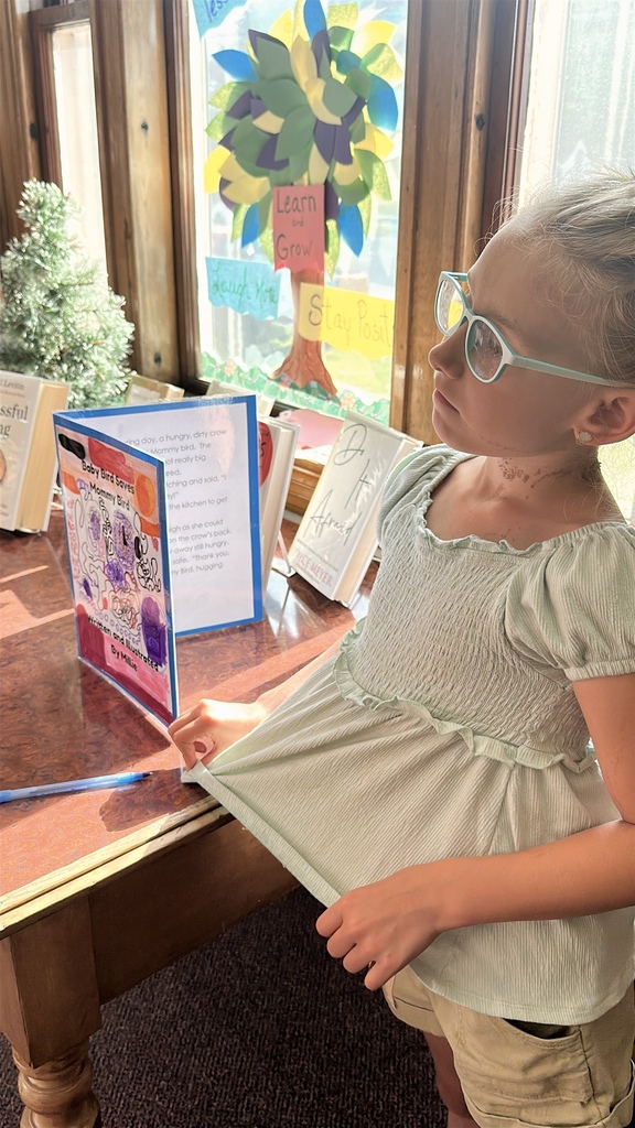 A student wearing glasses stands beside a display table, looking thoughtfully at a handmade and illustrated student book displayed in the library.