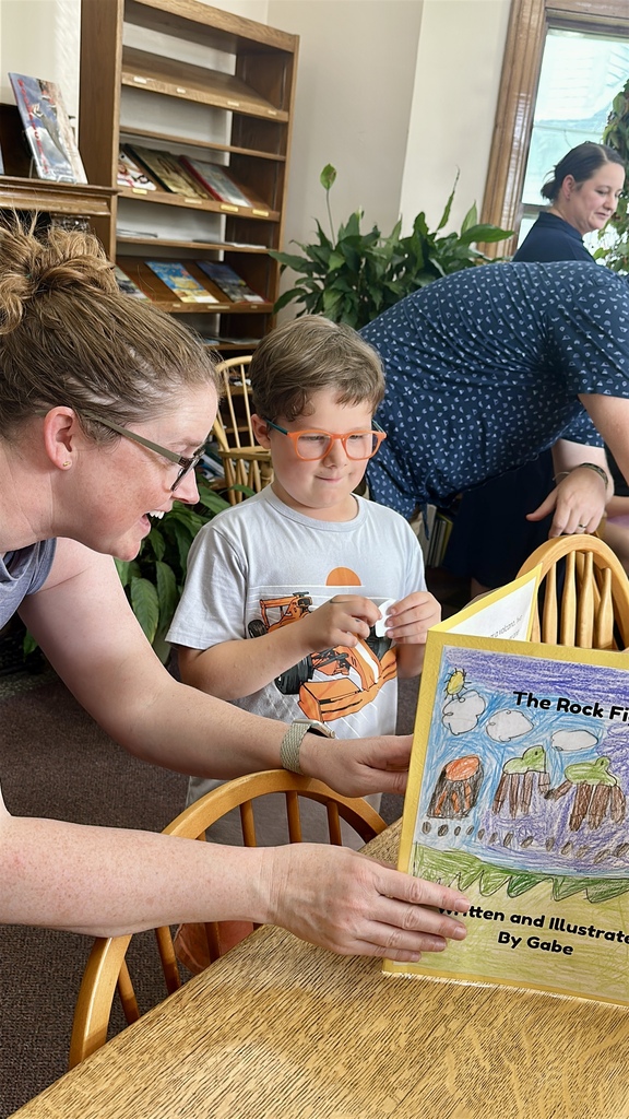 A student wearing orange glasses stands beside an adult while proudly sharing a colorful illustrated storybook during the book launch event.