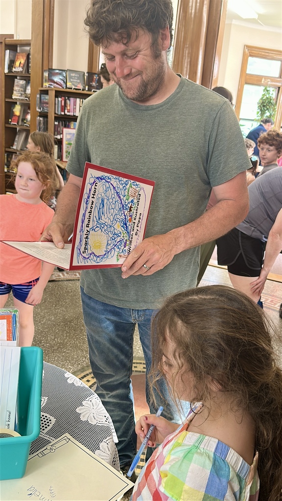 A smiling adult looks through a student's handmade storybook during the FHGS first grade book launch at the Fair Haven Free Library while children and families gather around displays of student work.