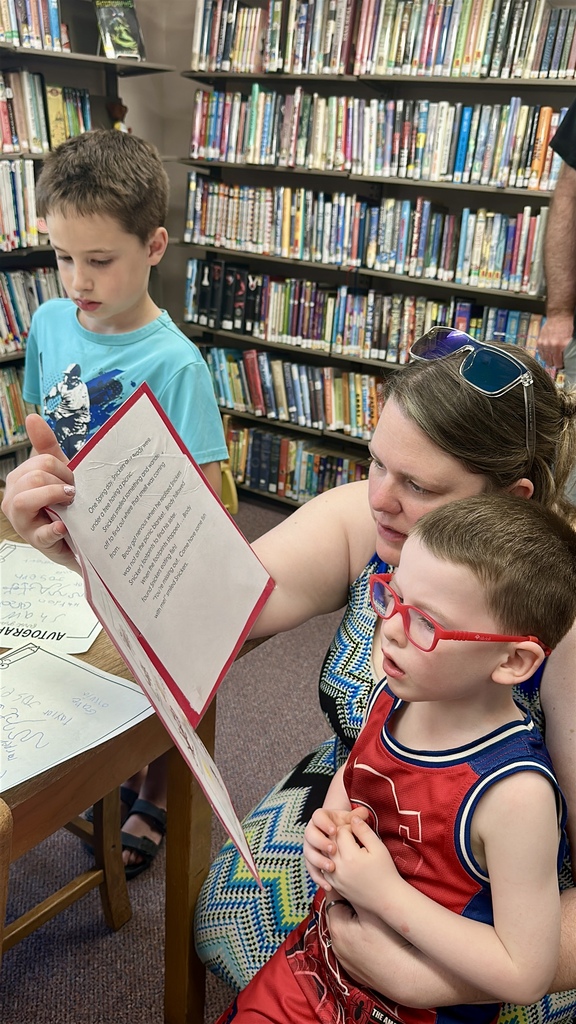 A parent and child sit together reading a student-created storybook at a table surrounded by library shelves during the FHGS book launch.