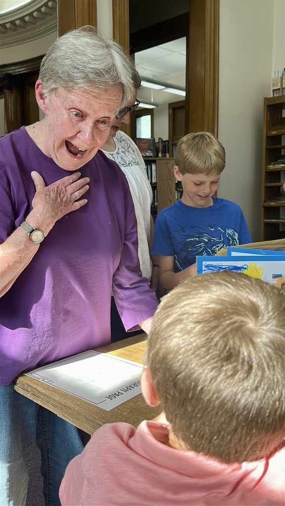 A family member reacts with excitement and surprise while looking through a student-created book as children gather around during the first grade author celebration.