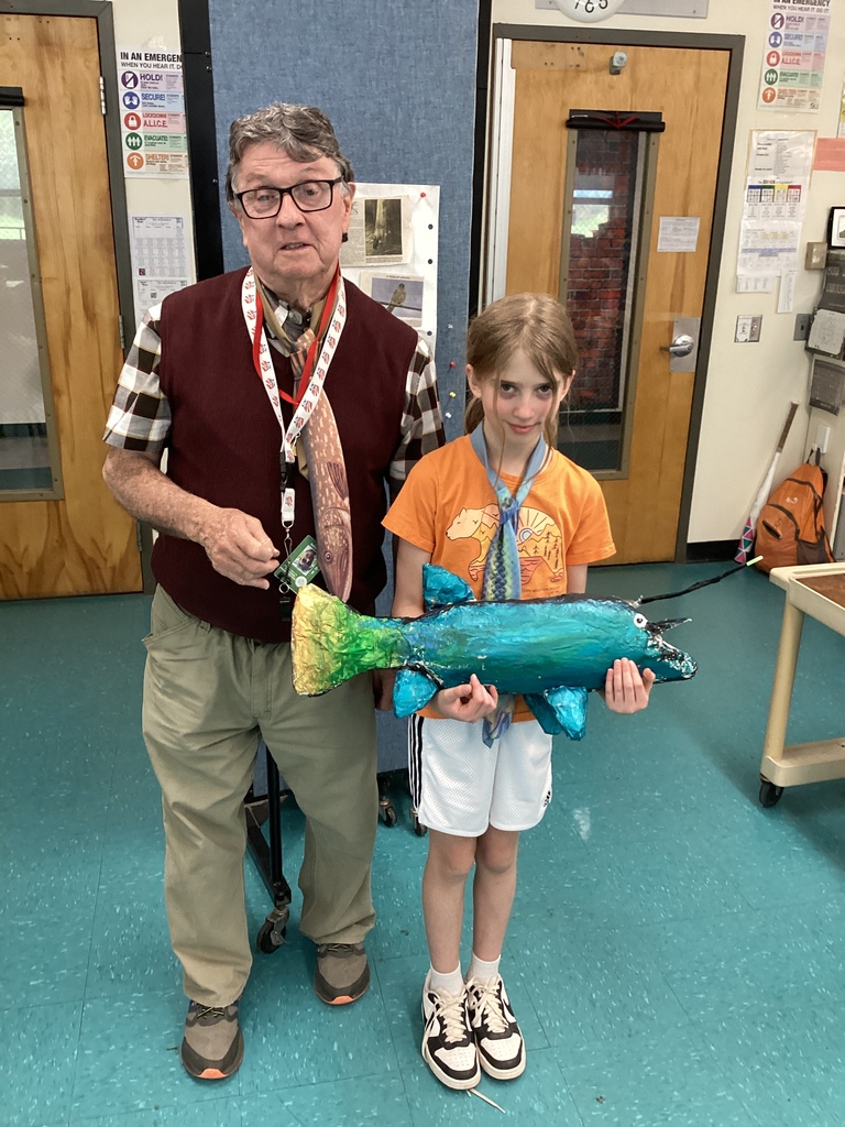 An older man and a student stand together in a classroom while the student holds a large handmade fish sculpture painted in shades of blue and green. Classroom doors, posters, and learning materials are visible in the background.