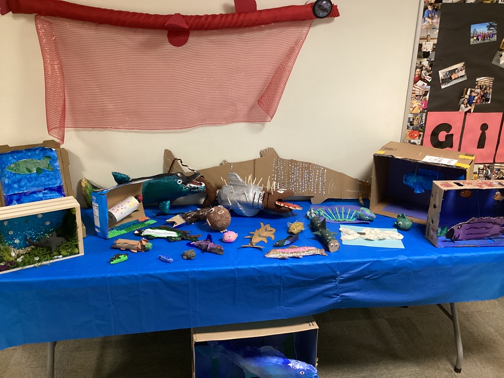 A display table covered with a blue tablecloth showcases a variety of student-created aquatic and fish-themed projects. Handmade fish sculptures, dioramas, and ocean-inspired creations are arranged across the table with decorative netting hanging above.