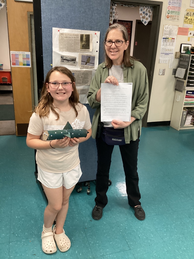A student and teacher pose together in a classroom displaying a fish project made from painted recycled materials along with a completed written assignment. Classroom posters and learning materials are visible in the background.
