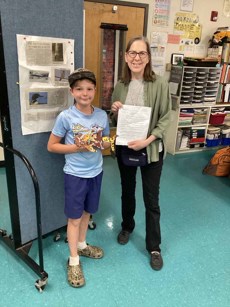 A student and teacher stand together in a classroom holding a written assignment and a colorful fish-themed project made from cut-out materials. A bulletin board with articles and classroom storage shelves are visible behind them.