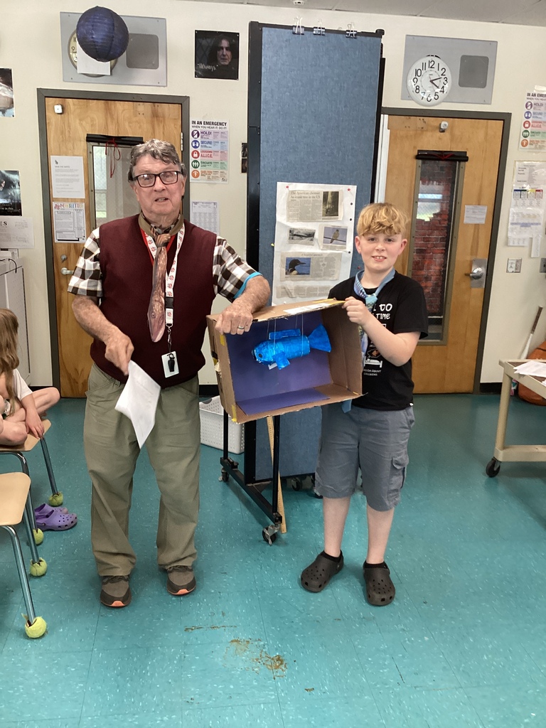 An older man and a student stand in a classroom displaying a student project inside a cardboard diorama featuring a hanging blue fish model. The student smiles while holding the project as classroom materials and posters appear in the background.