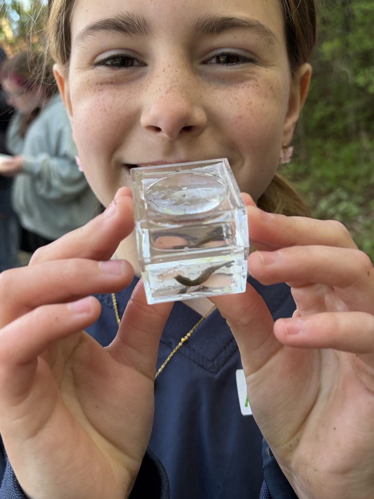 A close-up shot of a young girl with freckles holding a small, clear acrylic viewing cube up to the camera. Inside the water-filled cube, a small, dark aquatic creature resembles a leech or worm. The girl is smiling proudly, with her nose and mouth framed directly behind the transparent container.  