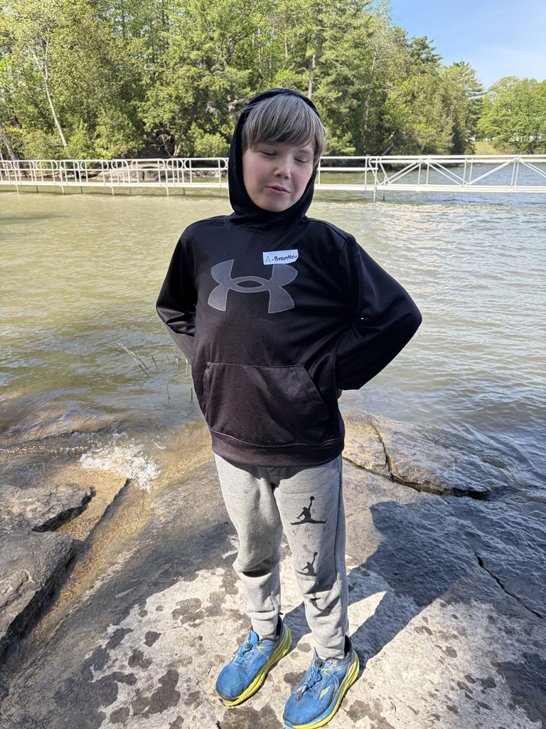  A full-length portrait of a young boy standing on a flat, rocky shoreline with water behind him. He wears a black Under Armour hooded sweatshirt with a small white name tag on the chest, grey Jordan brand sweatpants, and bright blue and yellow athletic shoes. He is smiling with his eyes closed and his hands resting behind his back. A long, white floating dock stretches across the water in the background beneath a treeline and blue sky.  