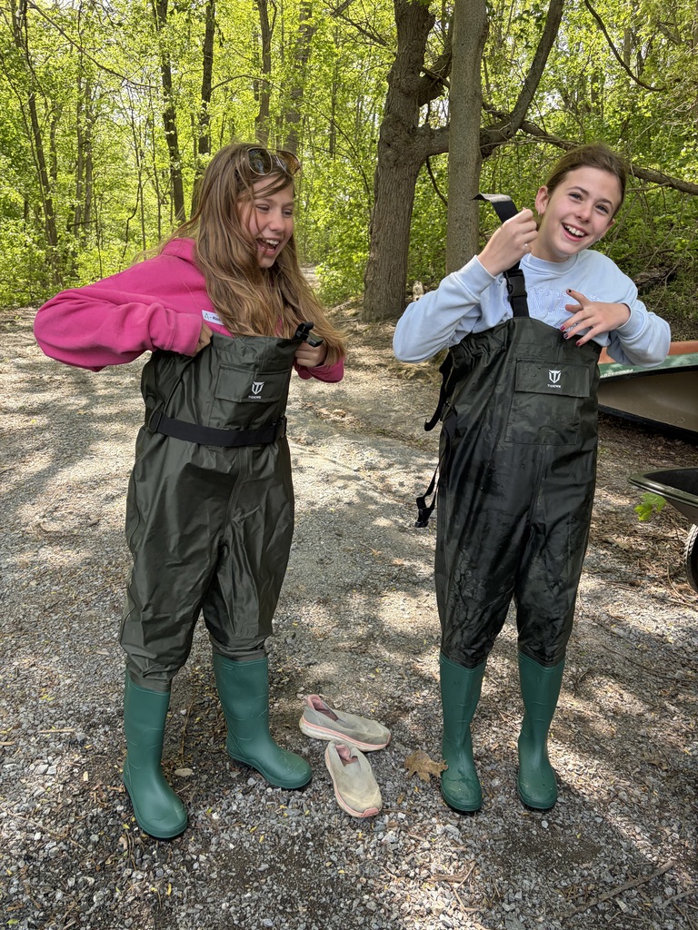 wo young girls stand side-by-side on a gravel path, laughing and smiling brightly as they struggle to pull up and adjust their oversized, baggy dark green chest waders. Their discarded sneakers sit on the ground between them, and a densely wooded area serves as the background. 
