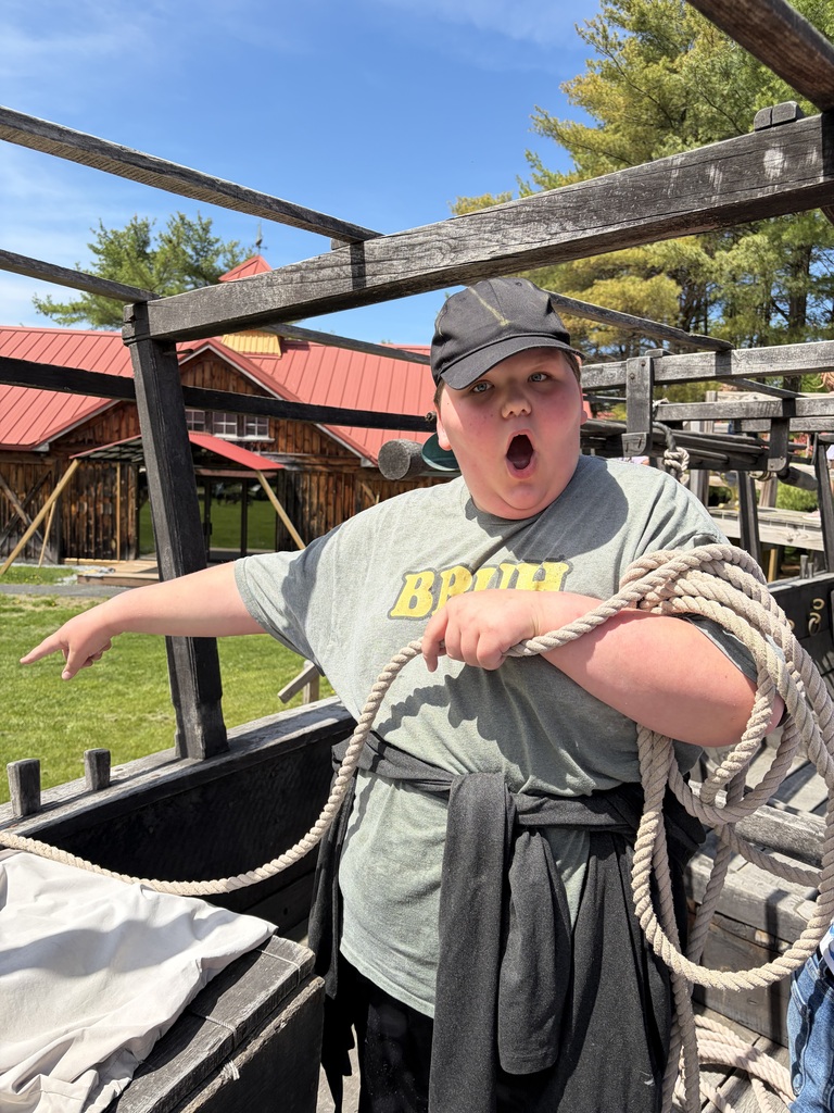 A medium shot of a young boy posing playfully inside the open wooden frame of what looks like a replica boat or structure. He is wearing a grey t-shirt that says "BRUH" in yellow letters, a black baseball cap, and has a black sweatshirt tied around his waist. He holds a thick, coiled beige rope over his shoulder, points off-camera to the left with an exaggerated, wide-open surprised expression on his face. A rustic wooden building with a red metal roof is visible in the background under a clear blue sky.  