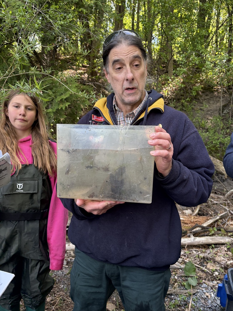 A close-up shot of an adult man with long dark hair pulled back and sunglasses resting on his head, standing in a wooded area. He is wearing a dark blue fleece jacket with a yellow lining and is holding a clear, rectangular acrylic tank filled with murky pond water, pointing out tiny aquatic insects swimming inside. To his left, a young girl in a pink hoodie and dark green waterproof chest waders stands watching him with a neutral expression. 