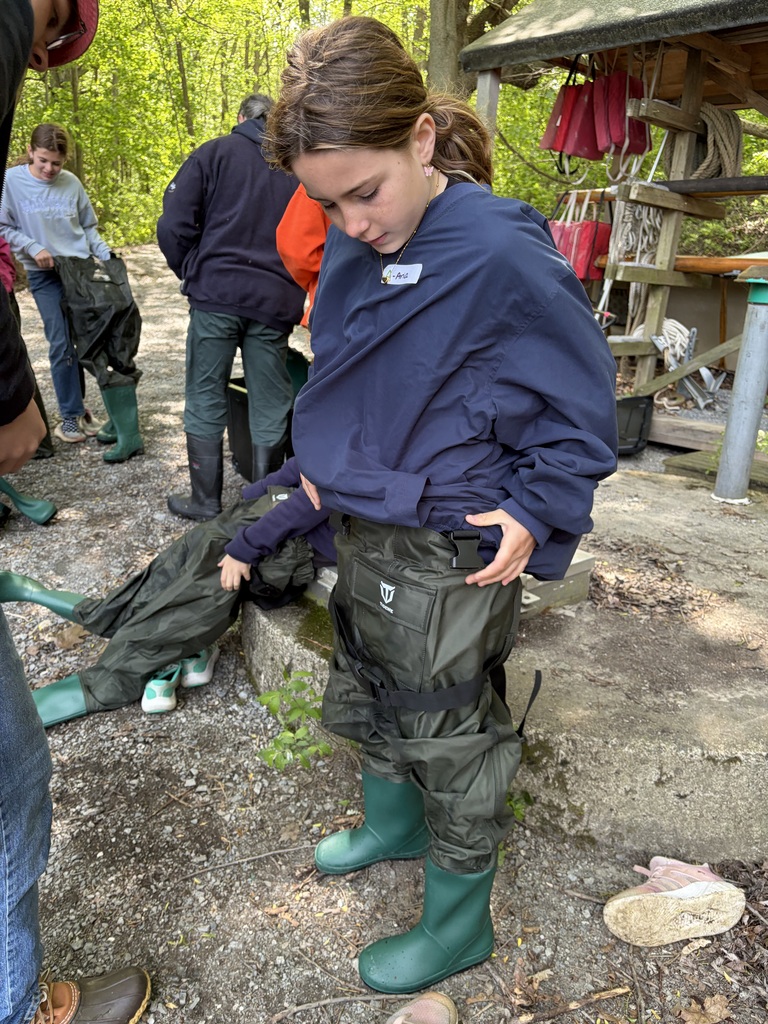  A young girl looks down as she pulls up a pair of oversized, dark green chest waders with integrated rubber boots. She is outdoors on a gravel path near a wooded area, surrounded by other people also gearing up. In the background, life jackets hang under a wooden shelter.