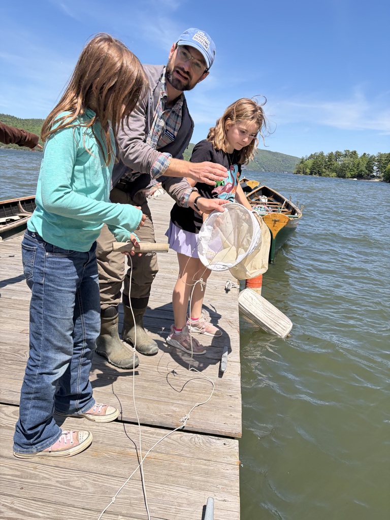 An outdoor shot on a sunny wooden dock where an instructor is teaching two young girls how to use a plankton net. The instructor, wearing a blue cap from the Lake Champlain Maritime Museum, leans forward to guide a white mesh cone-shaped net attached to a rope over the side of the dock into the water. One girl in a teal long-sleeve shirt and jeans holds a wooden handle attached to the line, while another girl in a black graphic t-shirt looks on intently. A green and yellow canoe is tied up to the