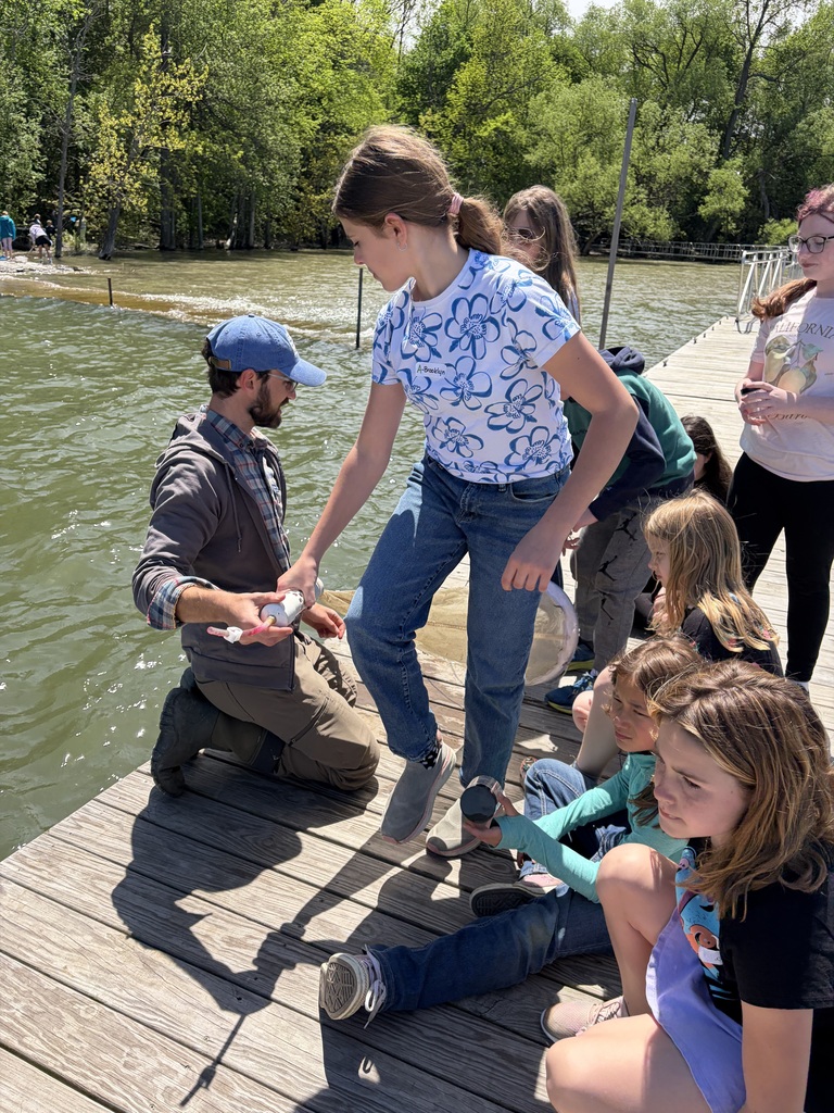 An outdoor shot on a wooden dock where a group of children are gathered around an instructor. The instructor, a man with a beard wearing a blue baseball cap, plaid shirt, and brown hoodie, kneels on the deck and holds out a small white collection vial. A young girl standing in a white t-shirt with large blue floral outlines and blue jeans leans forward to inspect it. Several other children sit or stand nearby on the dock under a bright, sunny sky, with green trees lining the distant shore