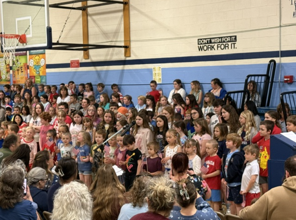 Students and staff gather together to sing during the Memorial Day program, with children wearing patriotic colors and families seated nearby.